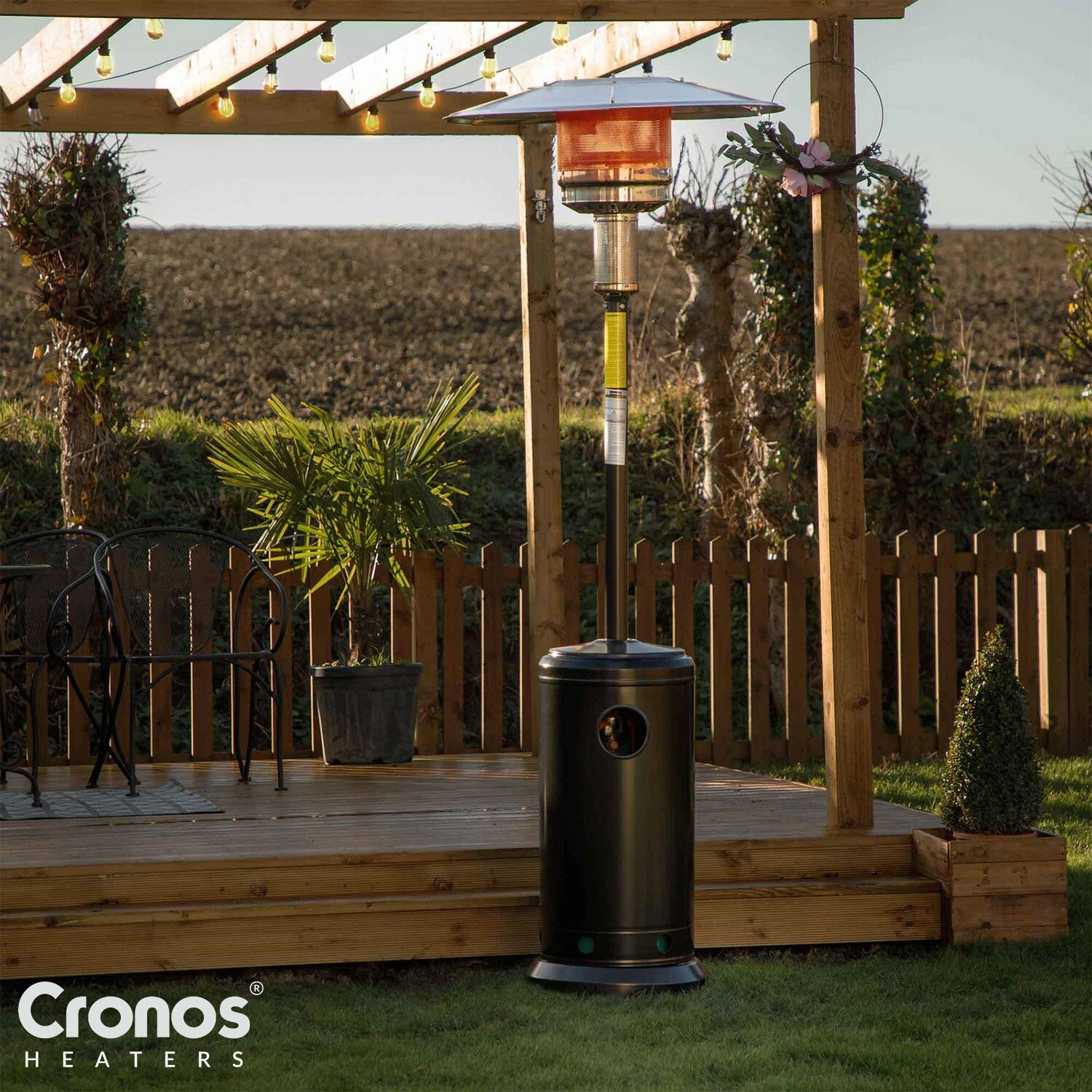Na drewnianym tarasie stoi czarny ogrzewacz tarasowy z metalowym daszkiem. W tle widoczny jest drewniany płot i rośliny, a w lewym dolnym rogu znajduje się logo \'Cronos Heaters\'.