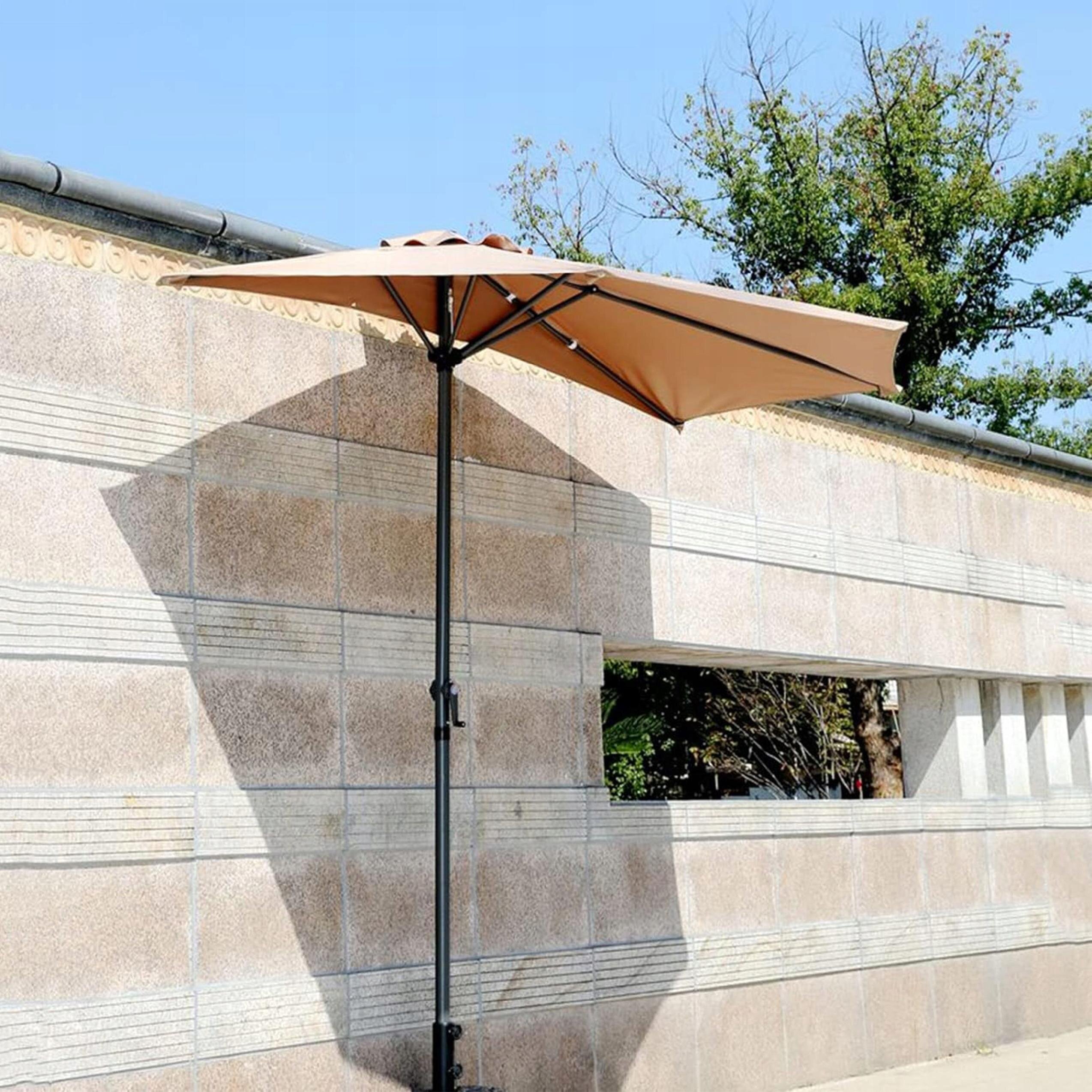 Duży beżowy parasol ogrodowy zamontowany przy ścianie, z metalowym stelażem i korbką do składania. W tle widoczna jest kamienna ściana z ozdobnymi wzorami i otworem, przez który widać drzewa.