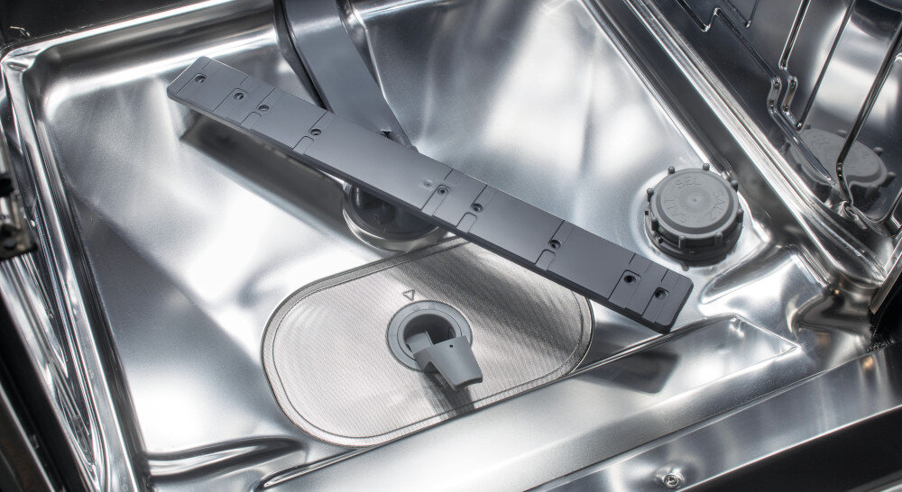 Wnętrze zmywarki z widocznym metalowym filtrem i plastikowym elementem w środku. W tle znajduje się okrągły element z tworzywa sztucznego.