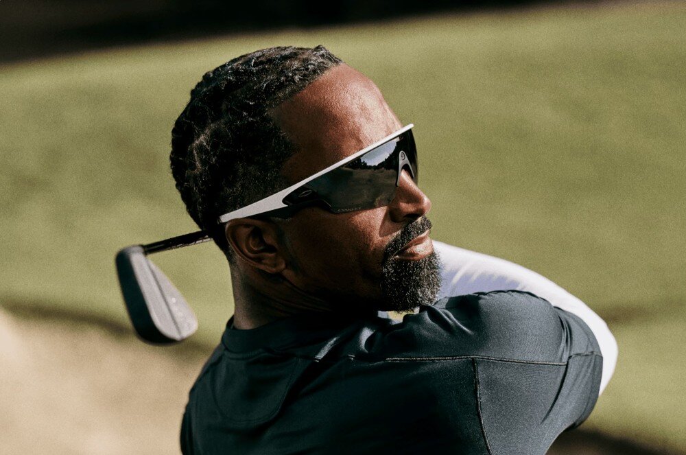 Okulary META Oakley Vanguad Mężczyzna z brodą w ciemnej koszulce i okularach przeciwsłonecznych trzyma kij golfowy na tle zielonej trawy, opis