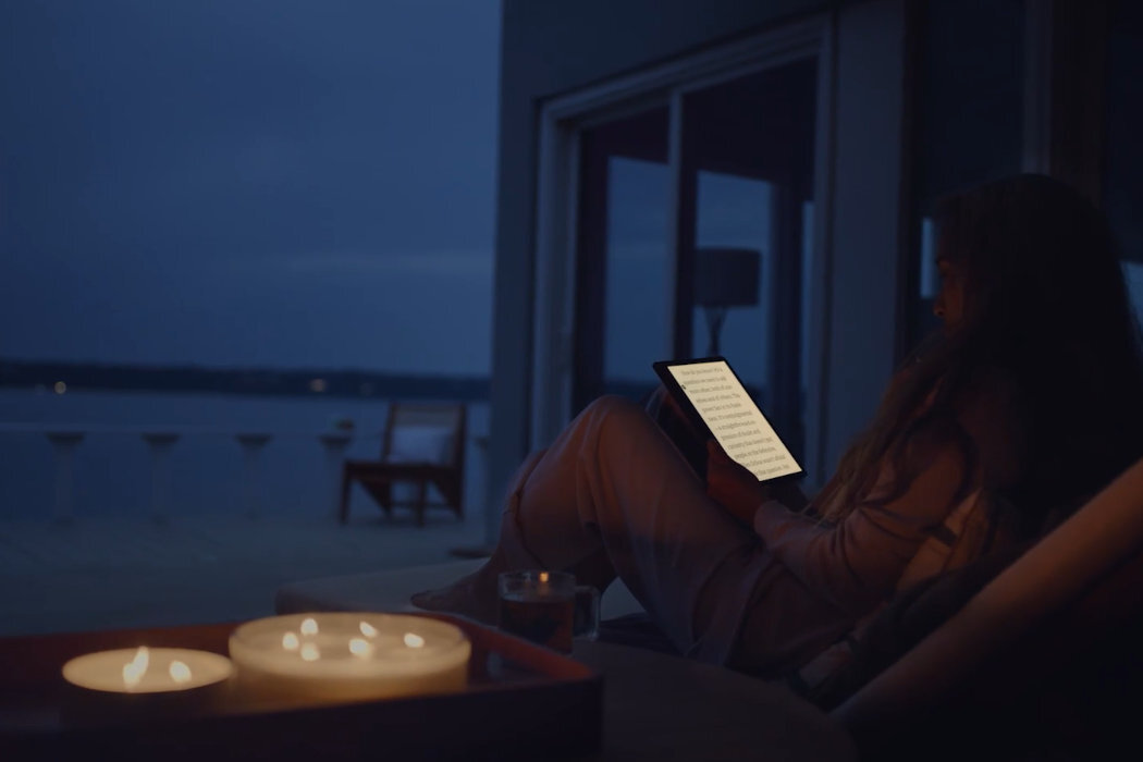 Osoba siedzi na tarasie w nocy, trzymając podświetlony czytnik e-booków. Na stoliku obok znajdują się zapalone świece.