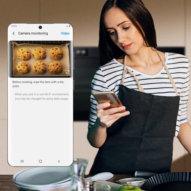 Kobieta w fartuchu trzyma telefon, na którym wyświetlany jest obrazek z piekarnika z piekącymi się ciasteczkami. Na ekranie telefonu widoczny jest tekst: \'Before cooking, wipe the lens with a dry cloth. - When you use in a non Wi-Fi environment, you may be charged for extra data usage.\'
