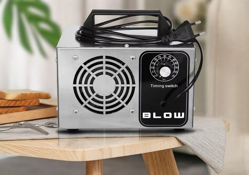 Na drewnianym stole stoi metalowy ozonator z widocznym wentylatorem i pokrętłem z napisem \'Timing switch\'. Na urządzeniu znajduje się logo \'BLOW\'.
