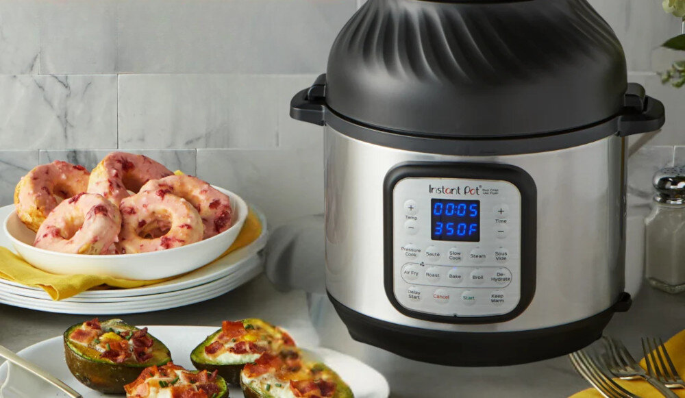 Na blacie kuchennym stoi multicooker z wyświetlaczem pokazującym \'00:05 350F\'. Obok znajdują się talerze z pączkami i zapiekanymi warzywami.