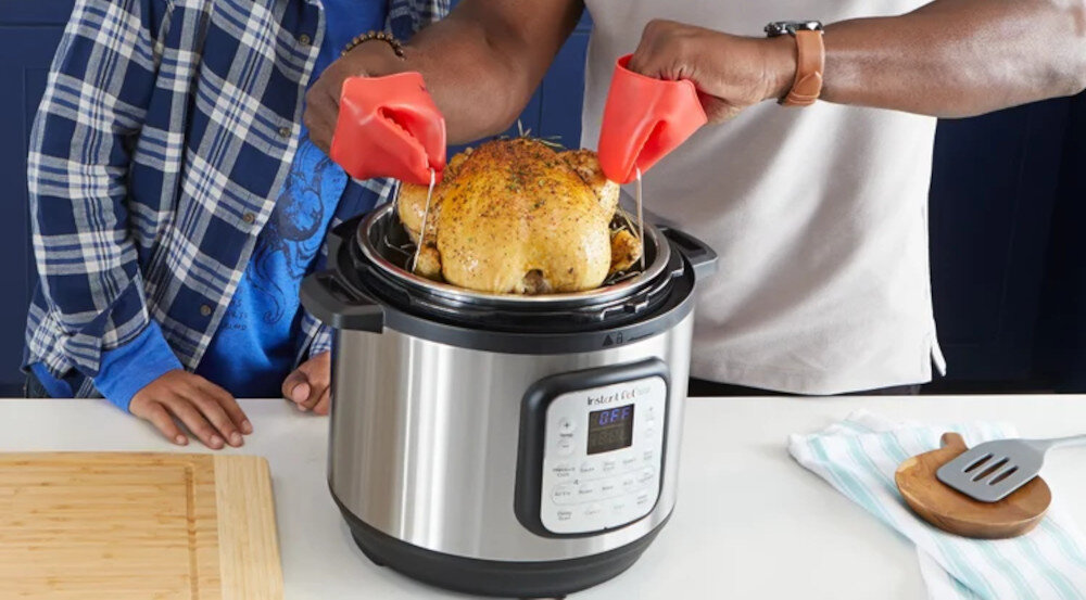 Osoba wyciąga pieczonego kurczaka z multicookera INSTANT POT przy użyciu czerwonych rękawic kuchennych. Na blacie obok znajduje się drewniana deska do krojenia i łopatka na ściereczce.