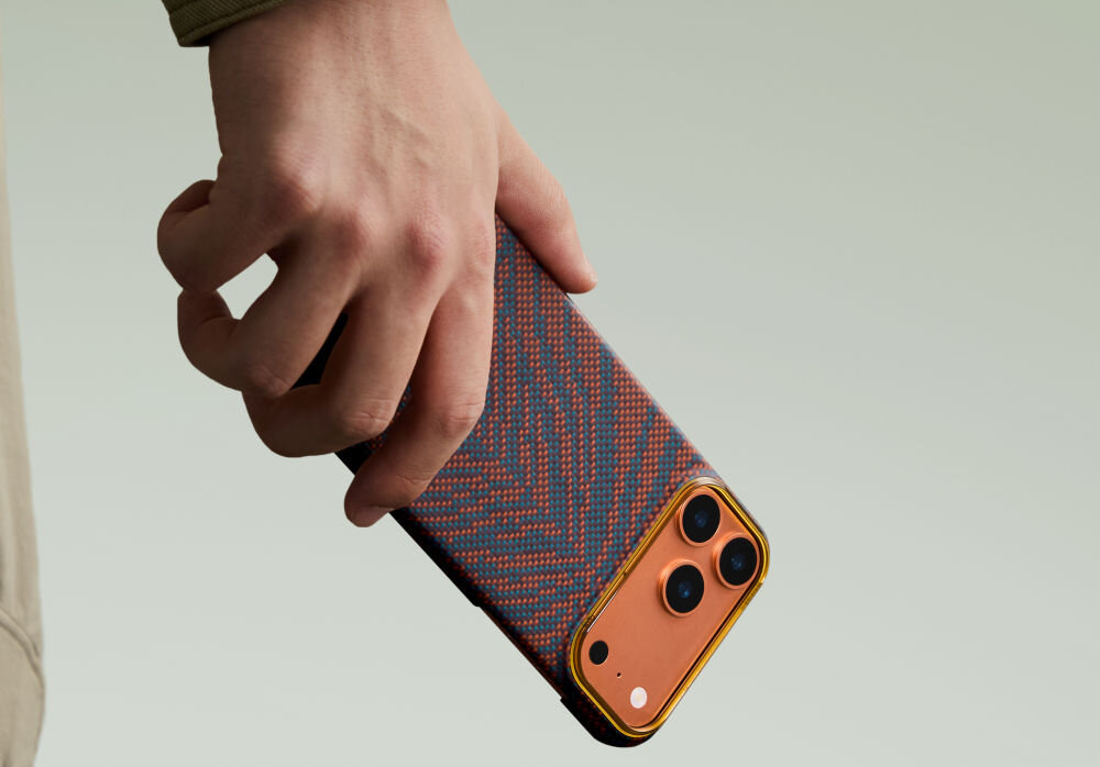 Osoba trzyma telefon w etui o wzorze w czerwono-niebieskie linie z pomarańczowym obramowaniem wokół aparatu. Tło jest jednolite i jasne.
