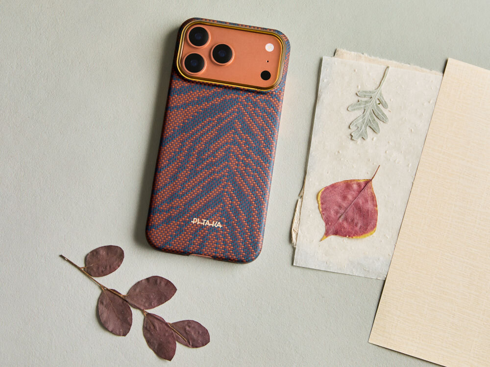 Na obrazie widoczne jest etui na telefon z wzorem w odcieniach pomarańczowego i niebieskiego, z napisem \'PITAKA\'. Obok leżą suszone liście i kartki papieru.