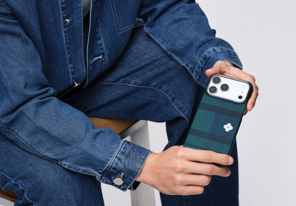 Osoba trzyma telefon w etui o wzorze kratki w odcieniach zieleni i granatu z białym symbolem kwiatka. Osoba ubrana jest w niebieski strój dżinsowy.