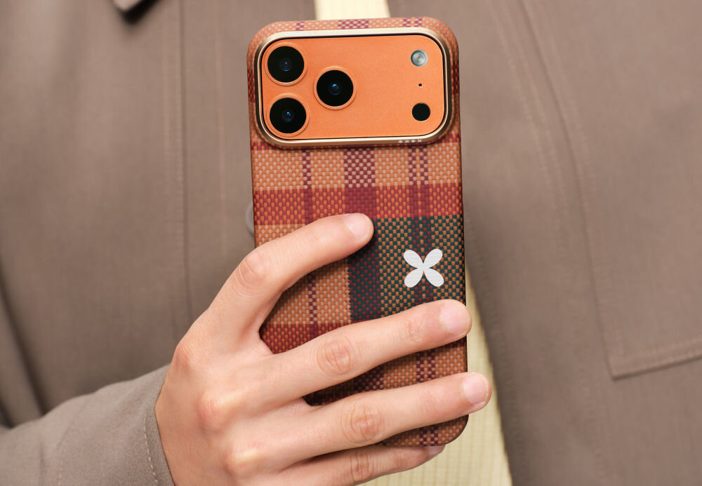 Osoba trzyma telefon w etui o wzorze w kratę z widocznym logo w kształcie białego kwiatu. Telefon ma pomarańczowy panel z trzema obiektywami aparatu.