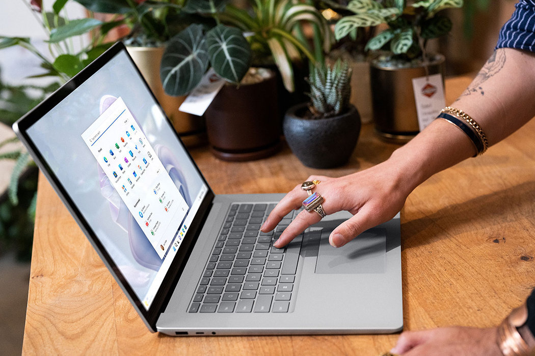 Osoba używa laptopa stojącego na drewnianym stole, a w tle widoczne są różne rośliny doniczkowe. Na ekranie laptopa wyświetlone są ikony aplikacji.