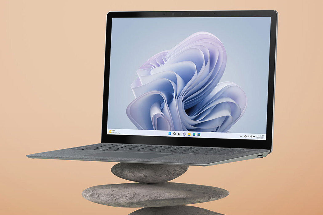 Laptop z ekranem wyświetlającym abstrakcyjną grafikę, umieszczony na stosie kamieni. Klawiatura wykonana z materiału przypominającego Alcantarę.