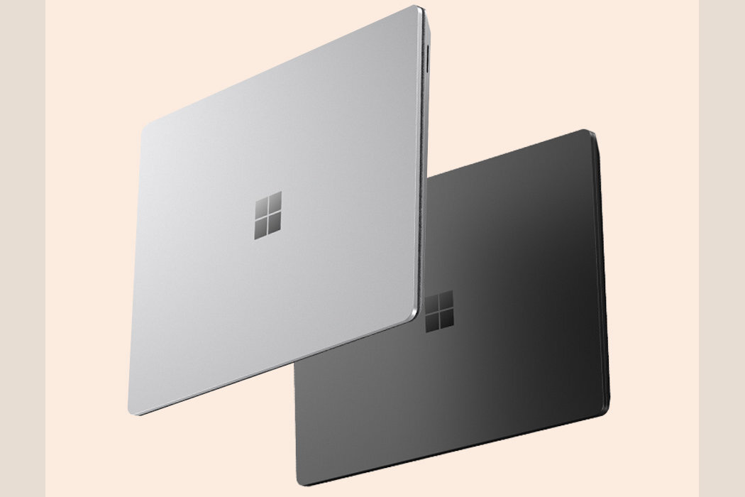 Dwa laptopy w kolorach srebrnym i ciemnoszarym, z widocznym logo w formie czterech kwadratów na pokrywie. Laptopy są pokazane w pozycji zamkniętej na jasnym tle.