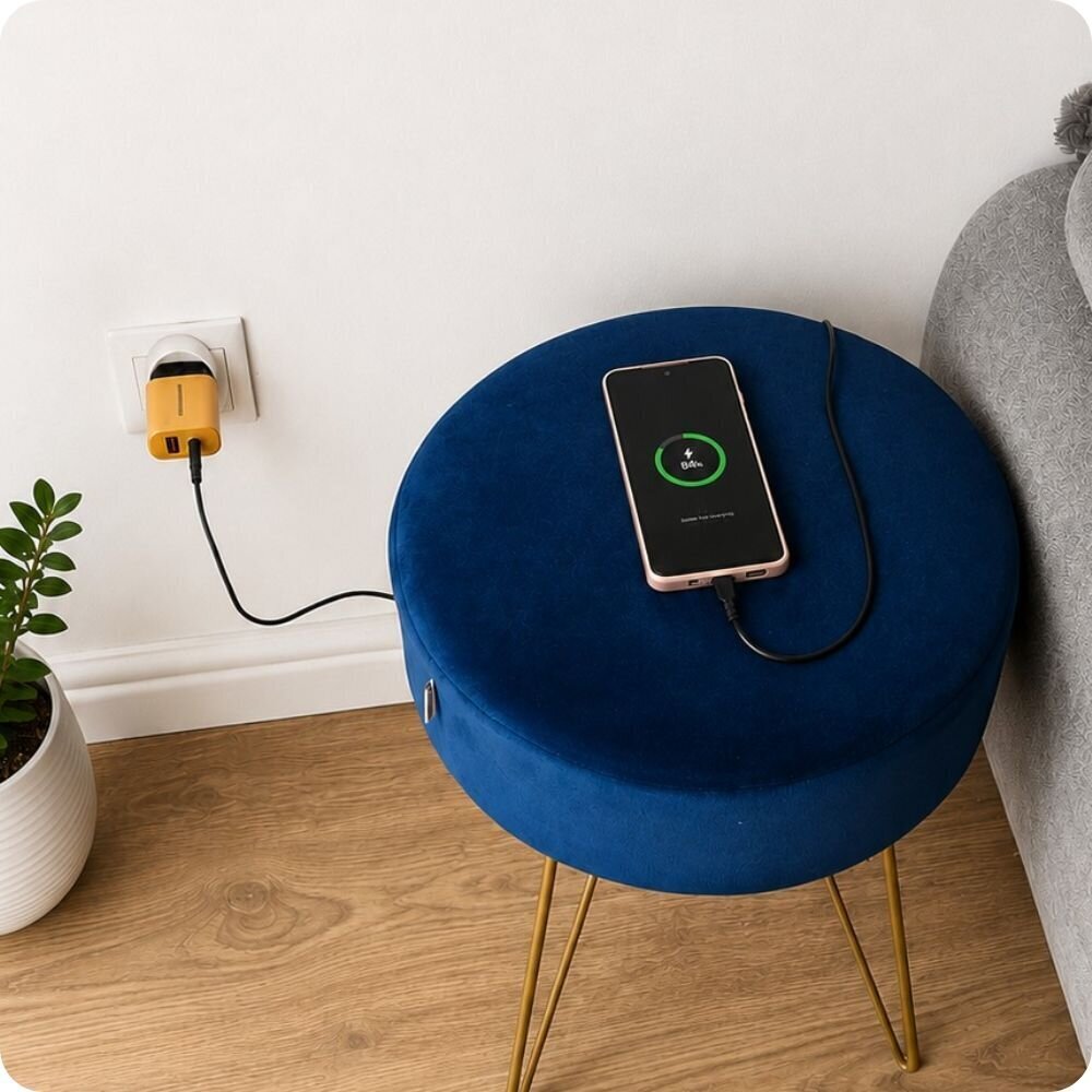 Telefon leży na niebieskim pufie, podłączony do ładowarki w gniazdku ściennym. Na ekranie telefonu widoczny jest symbol ładowania i napis \'84%\'.