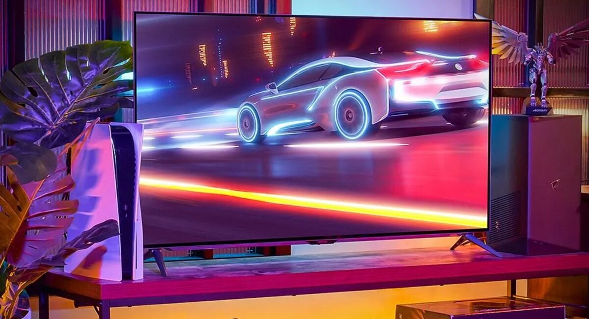 Na obrazie widać duży monitor wyświetlający dynamiczną scenę z futurystycznym samochodem w neonowych kolorach. Obok monitora znajduje się konsola do gier oraz dekoracyjna figurka.