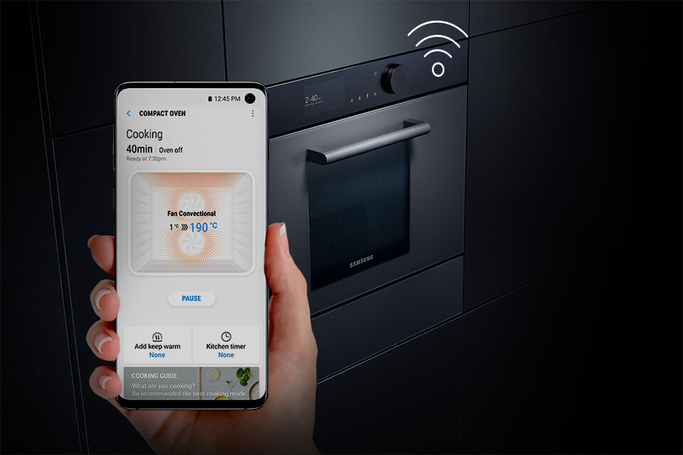 Osoba trzyma smartfon z aplikacją pokazującą ustawienia piekarnika, w tle widać wbudowany piekarnik z logo Samsung. Na ekranie telefonu widoczny jest tekst: \'COMPACT OVEN\', \'Cooking 40min Oven off\', \'Fan Convection 190°C\', \'PAUSE\', \'Add keep warm\', \'Kitchen timer\'.