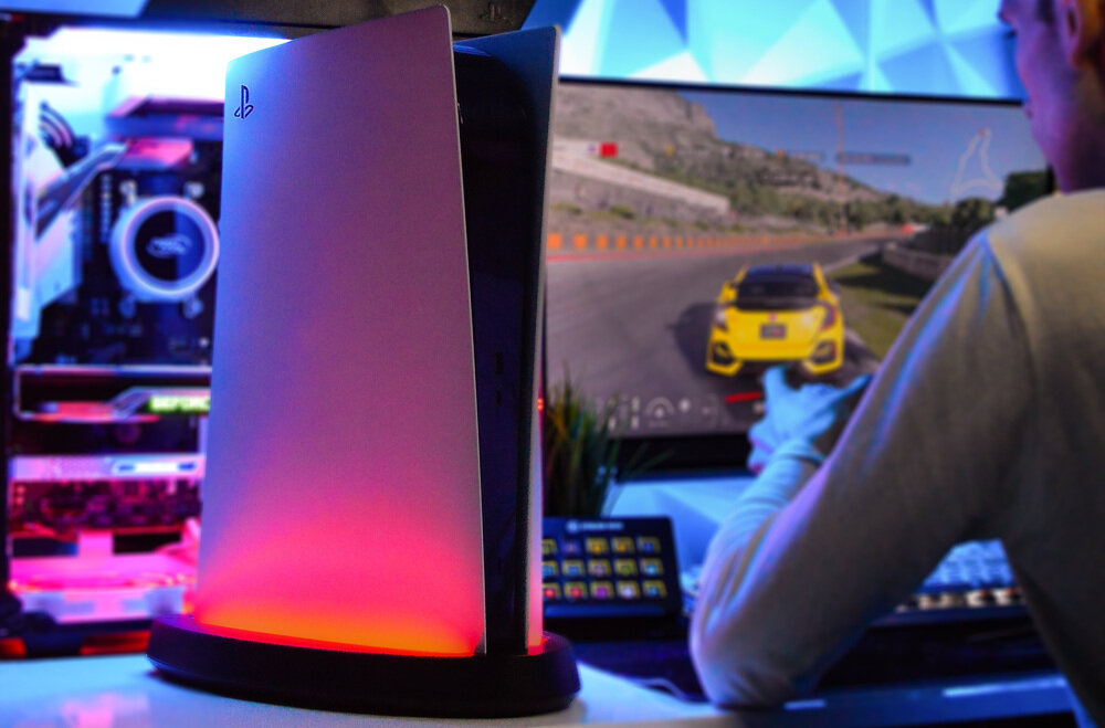 Konsola PlayStation 5 stoi na podświetlanej podstawce LED, emitującej czerwone światło. W tle widoczny jest monitor z grą wyścigową oraz osoba trzymająca kontroler.
