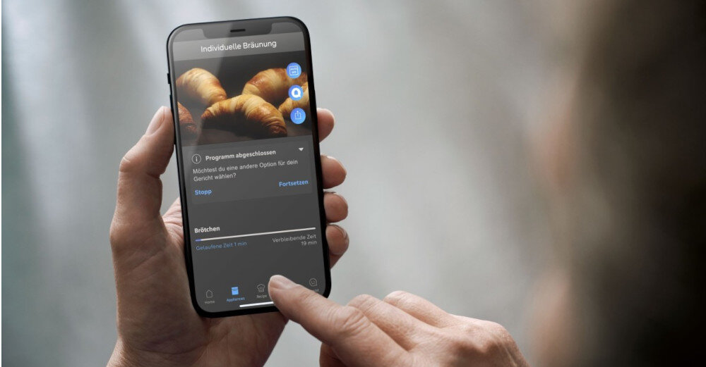 Osoba trzyma smartfon, na ekranie którego widoczna jest aplikacja z obrazem rogalików i tekstem \'Individuelle Bräunung\'.