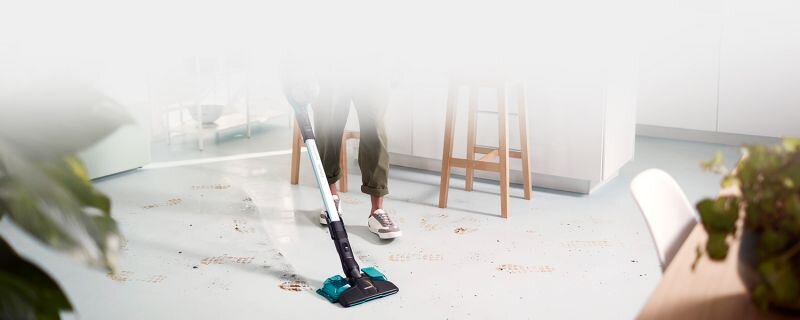 Osoba używa odkurzacza pionowego z funkcją mopowania na jasnej podłodze z widocznymi zabrudzeniami. W tle znajduje się kuchnia z białymi szafkami i krzesłami.