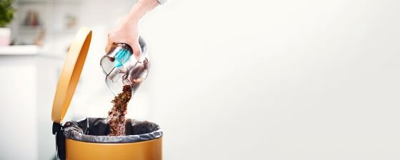 Osoba opróżnia pojemnik z odkurzacza do kosza na śmieci. Kosz ma pomarańczową pokrywę i czarny worek na śmieci.