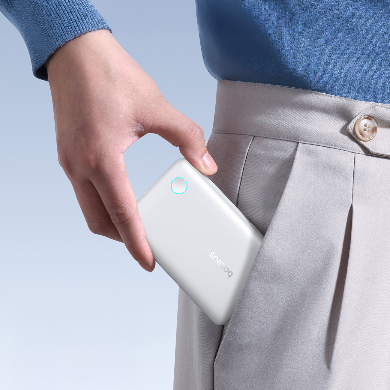 Osoba wkłada biały power bank z napisem \'Baseus\' do kieszeni jasnych spodni. Power bank ma okrągły, podświetlony przycisk.