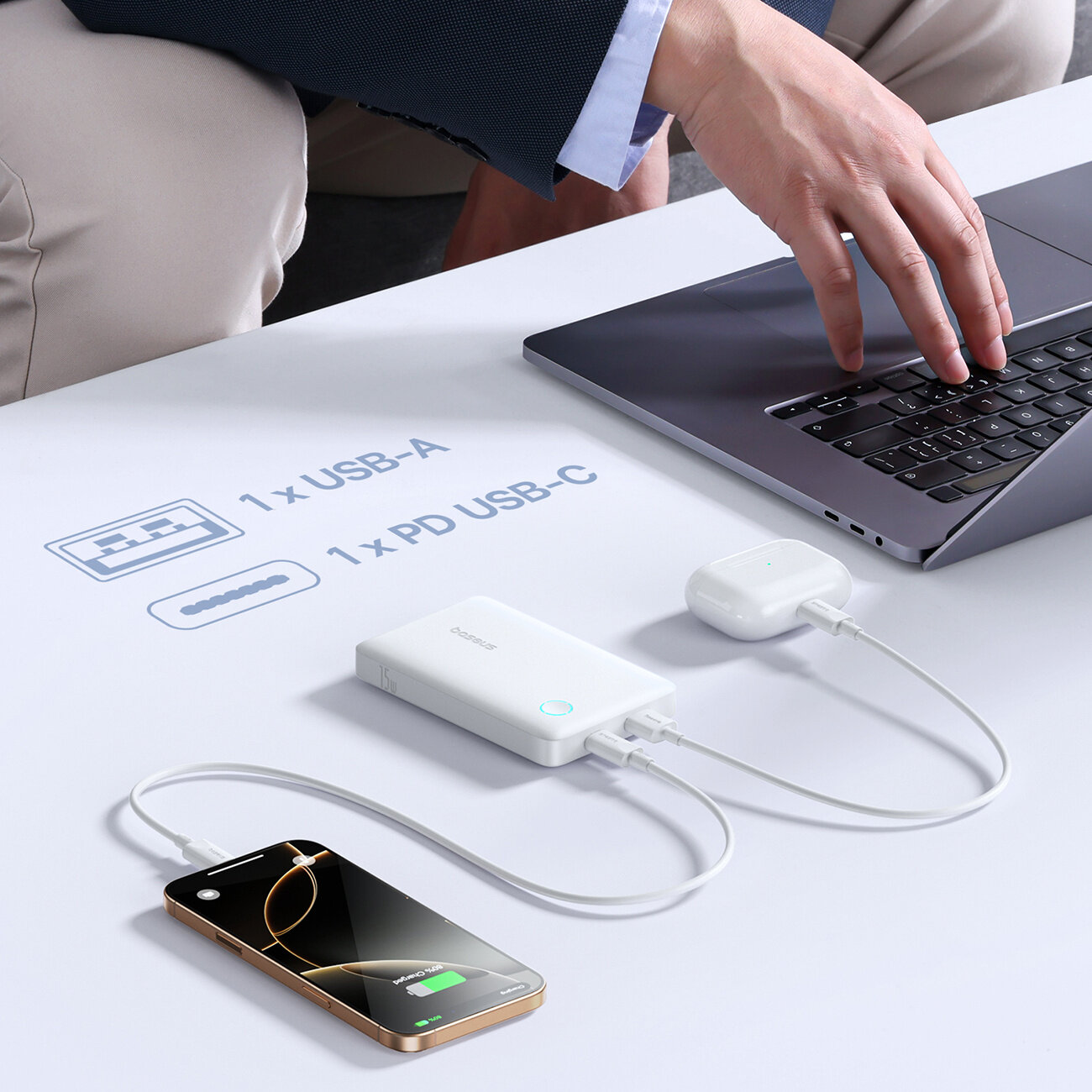 Na stole znajduje się biały power bank z logo \'Baseus\', do którego podłączone są dwa urządzenia: telefon i słuchawki. Obok widoczny jest laptop oraz tekst: \'1 x USB-A, 1 x PD USB-C\'.