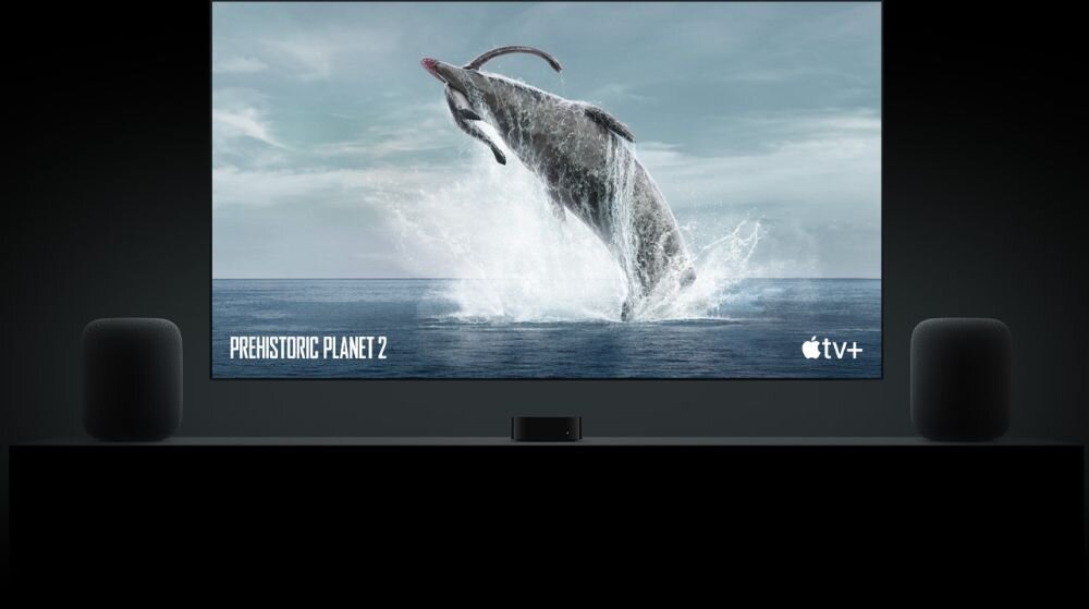 Na ekranie telewizora wyświetlany jest obraz skaczącego wieloryba z napisem \'PREHISTORIC PLANET 2\' oraz logo \'apple tv+\'. Po obu stronach telewizora znajdują się dwa czarne głośniki.