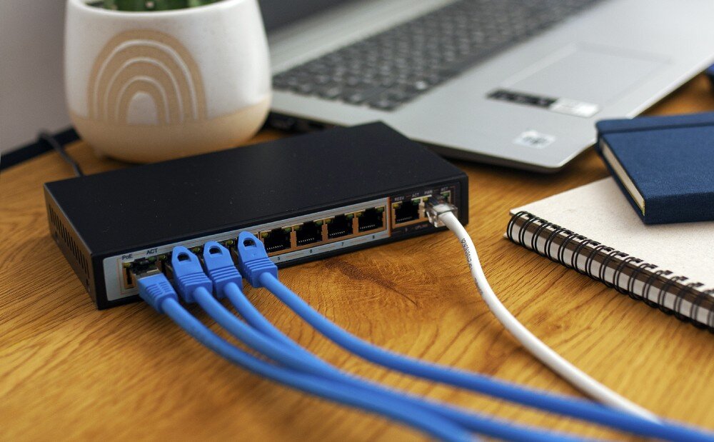 Switch EXTRALINK Ceres Czarny switch leżący na drewnianym biurku, z wpiętymi czterema niebieskimi i jednym białym kablem sieciowym, w tle laptop i notatnik