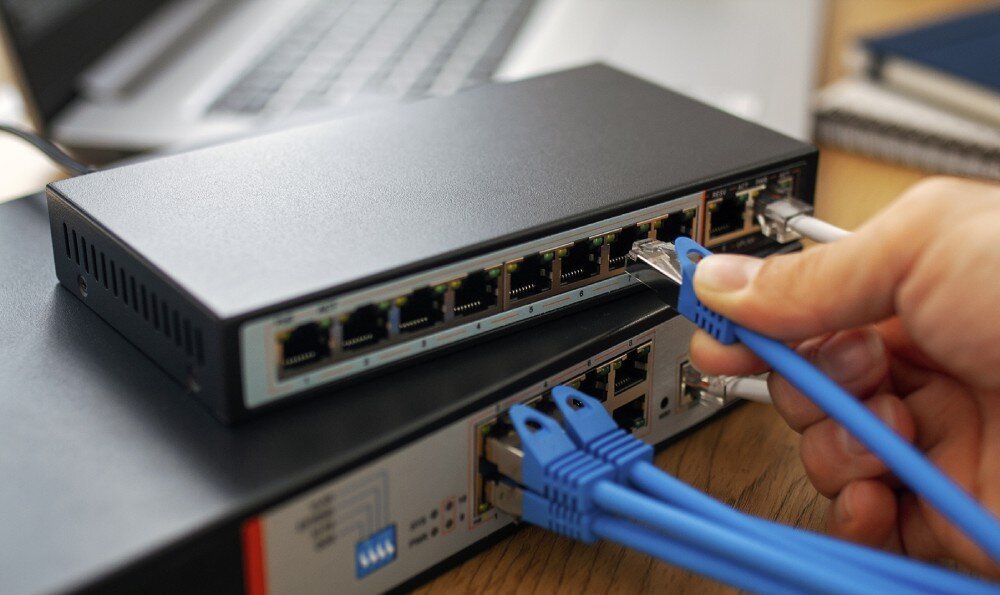 Switch EXTRALINK Ceres Dłoń wpinająca niebieski kabel sieciowy do czarnego switcha, który leży na innym, większym switchu