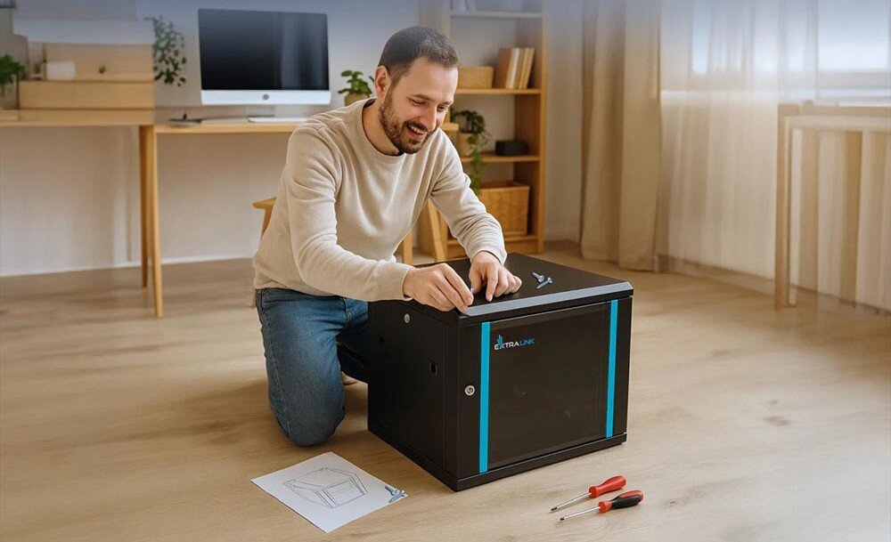 Szafa RACK EXTRALINK 19 4U EX.2893 600 x 600 mm Uśmiechnięty mężczyzna klęczący na podłodze podczas montażu czarnej szafki, obok leżą śrubokręty i instrukcja