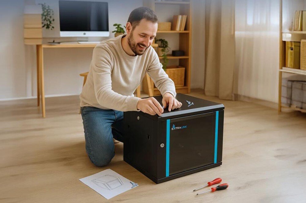 Szafa RACK EXTRALINK 19 6U EX.7232 600 x 600 mm Uśmiechnięty mężczyzna klęczący na podłodze podczas montażu czarnej szafki, obok leżą śrubokręty i instrukcja