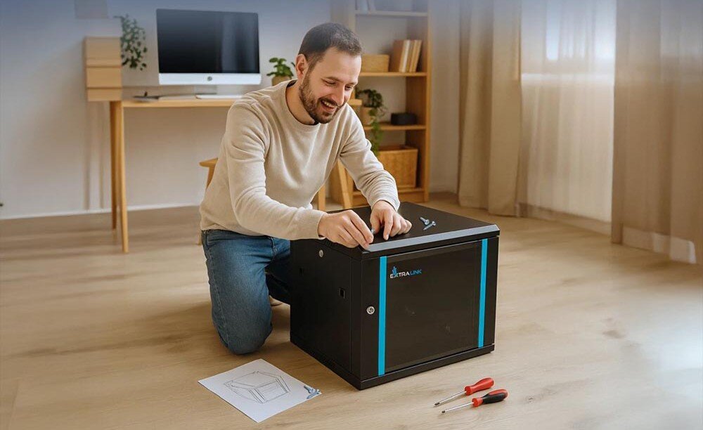 Szafa RACK EXTRALINK 19 9U EX.7249 600 x 450 mm Uśmiechnięty mężczyzna siedzący na podłodze i składający szafę serwerową, obok niego leżą śrubokręty i instrukcja, boczne panele