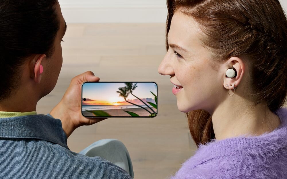 Osoba trzyma smartfon z wyświetlonym obrazem plaży o zachodzie słońca, obok inna osoba nosi bezprzewodowe słuchawki.