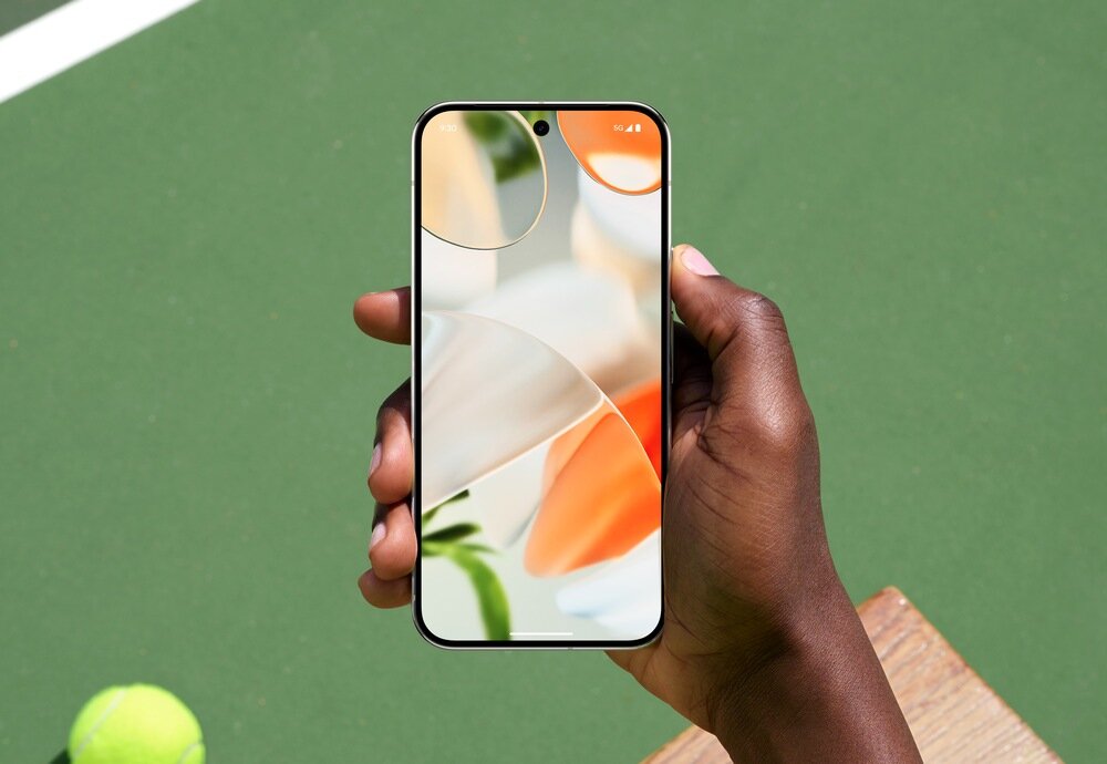 Osoba trzyma smartfon z wyświetlaczem pokazującym abstrakcyjną grafikę w jasnych kolorach. W tle widoczna jest zielona powierzchnia i piłka tenisowa.
