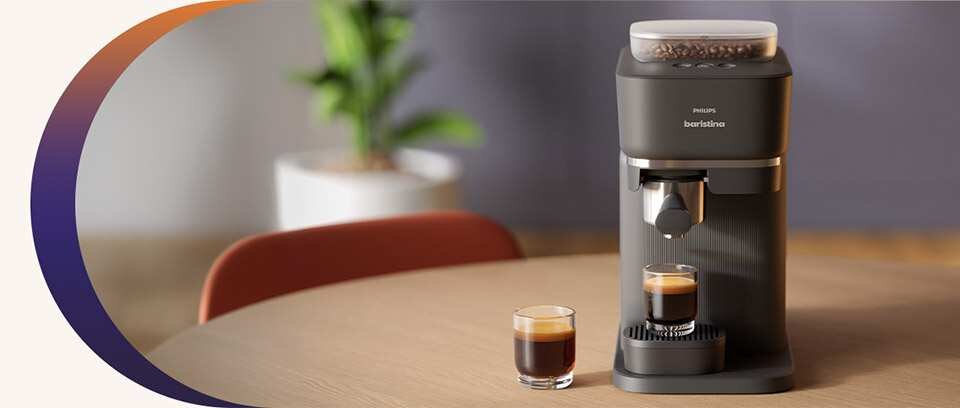 Na drewnianym stole stoi ekspres do kawy z logo \'PHILIPS baristina\', obok którego znajdują się dwie filiżanki z kawą. W tle widoczna jest rozmyta roślina w doniczce.