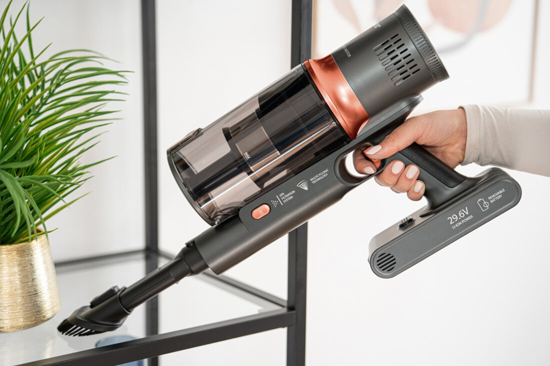 Osoba trzyma odkurzacz ręczny z widocznym napisem \'29.6V li-ion power\'. W tle znajduje się zielona roślina w złotej doniczce.