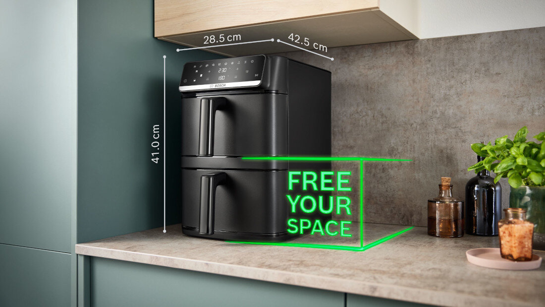 Na blacie kuchennym stoi czarna frytkownica beztłuszczowa z dwoma koszami, obok której widnieje napis \'FREE YOUR SPACE\'. Wymiary urządzenia to 28.5 cm szerokości, 42.5 cm głębokości i 41.0 cm wysokości.