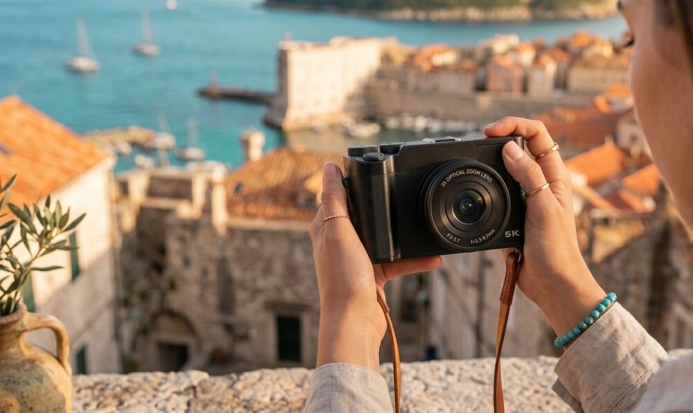 Osoba trzyma aparat fotograficzny z widocznym napisem \'OPTICAL ZOOM LENS 5X\' na tle zabytkowego miasta i morza.