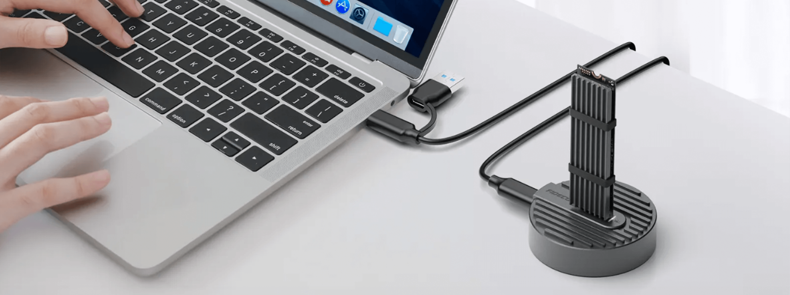 Na biurku znajduje się otwarty laptop z podłączonym adapterem SSD, który stoi pionowo na okrągłej podstawce. Obok laptopa widoczne są ręce osoby korzystającej z touchpada.
