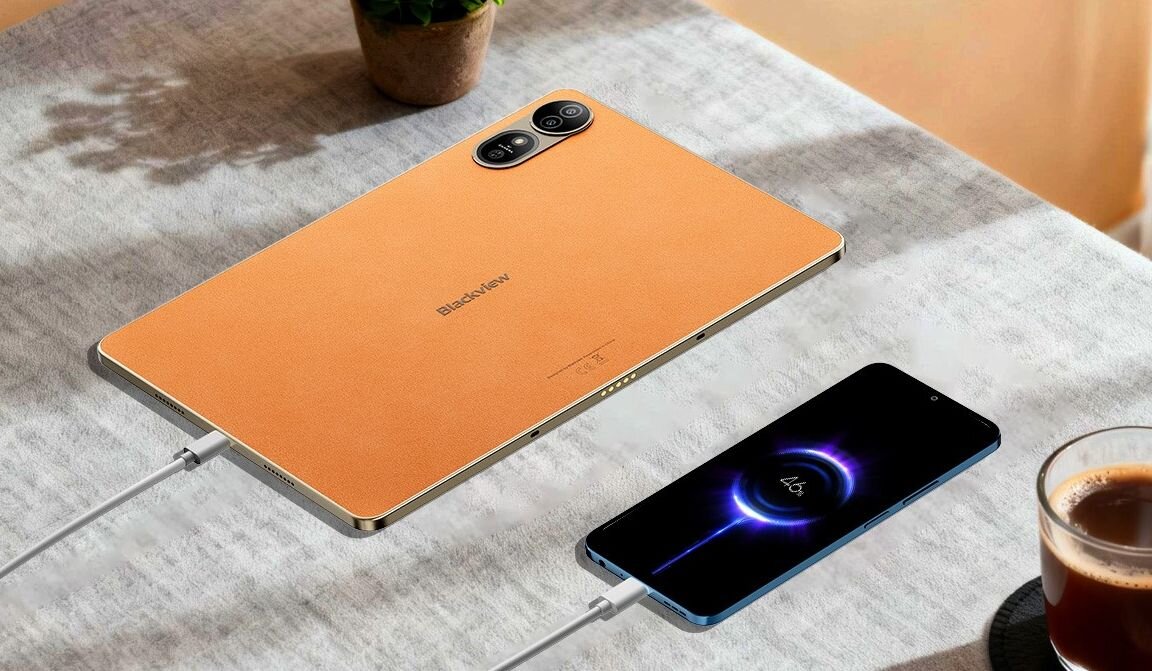 Na stole leży tablet w kolorze pomarańczowym z widocznym logo \'Blackview\' oraz smartfon z wyświetlonym symbolem ładowania. Oba urządzenia są podłączone do ładowania, a obok znajduje się filiżanka z napojem.