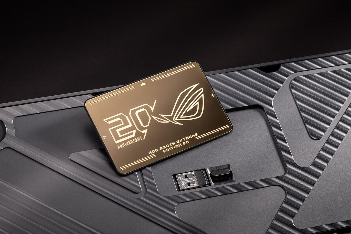 Na obrazie widoczna jest metalowa tabliczka z napisem \'20th Anniversary ROG Azoth Extreme Edition 20\' oraz logo ROG. Tabliczka leży na powierzchni o teksturze przypominającej włókno węglowe.