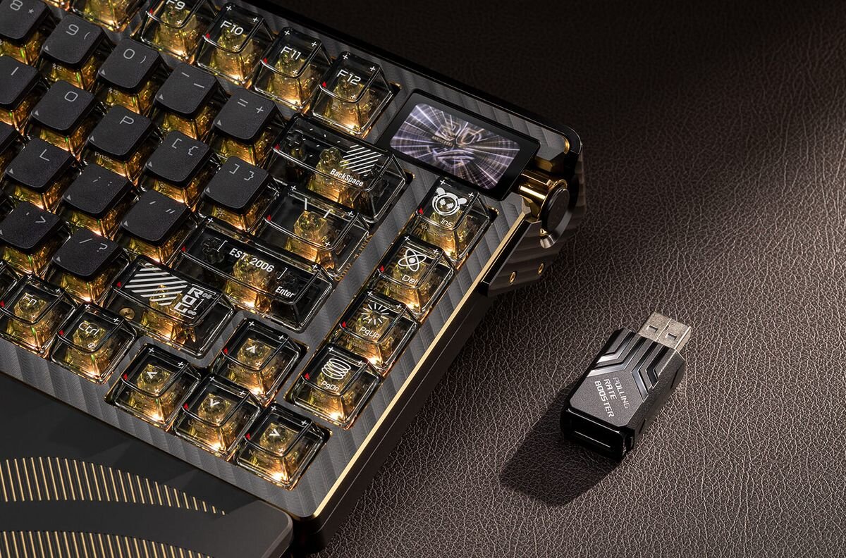 Na obrazie widoczna jest klawiatura z podświetlanymi klawiszami oraz mały wyświetlacz z grafiką. Obok klawiatury leży adapter USB z napisem \'ROG STRIX\'.