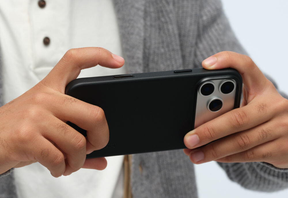 Osoba trzyma czarny telefon w etui, widoczne są trzy obiektywy aparatu. Telefon jest trzymany obiema rękami, a w tle widać fragment szarego swetra i białej koszuli.
