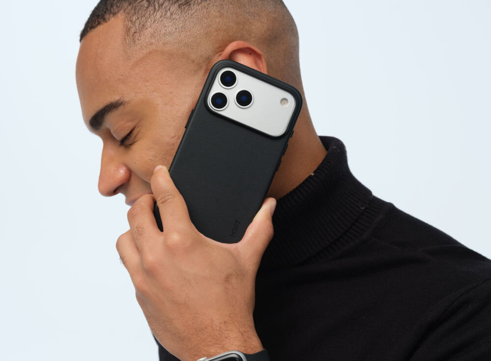 Osoba trzyma przy uchu telefon w czarnym etui z trzema widocznymi obiektywami aparatu. Na etui widoczny jest napis \'MOFT\'.