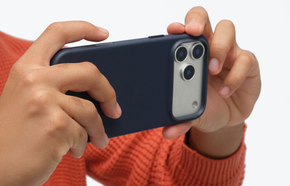Osoba trzyma telefon w granatowym etui, widoczne są trzy obiektywy aparatu. Osoba ma na sobie pomarańczowy sweter.