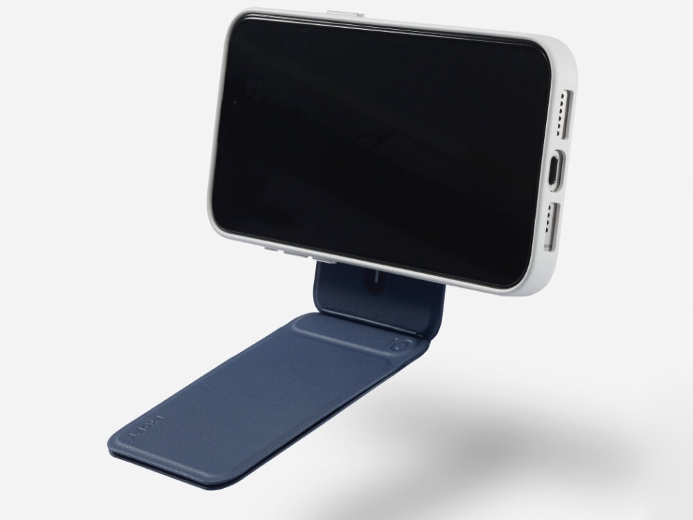 Granatowy stojak na telefon z funkcją MagSafe, na którym zamocowany jest biały telefon. Na stojaku widoczne jest logo \'MOFT\'.