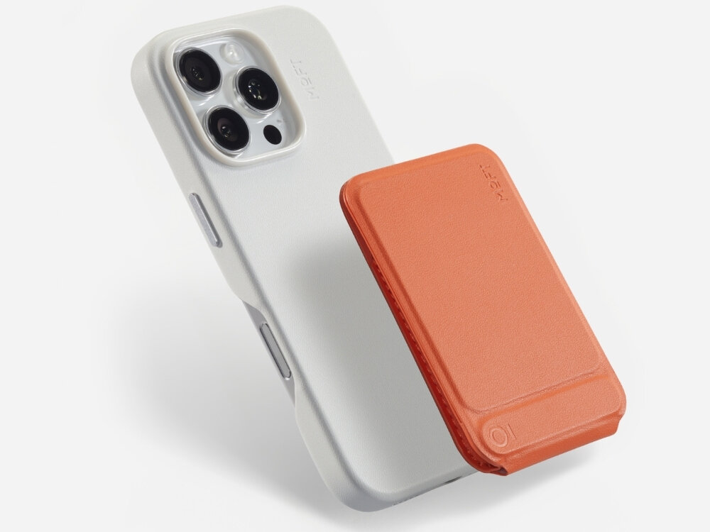 Na obrazie widoczny jest biały telefon z trzema obiektywami aparatu oraz pomarańczowy stojak magnetyczny przymocowany do jego tylnej części. Na stojaku widnieje napis \'MOFT\'.
