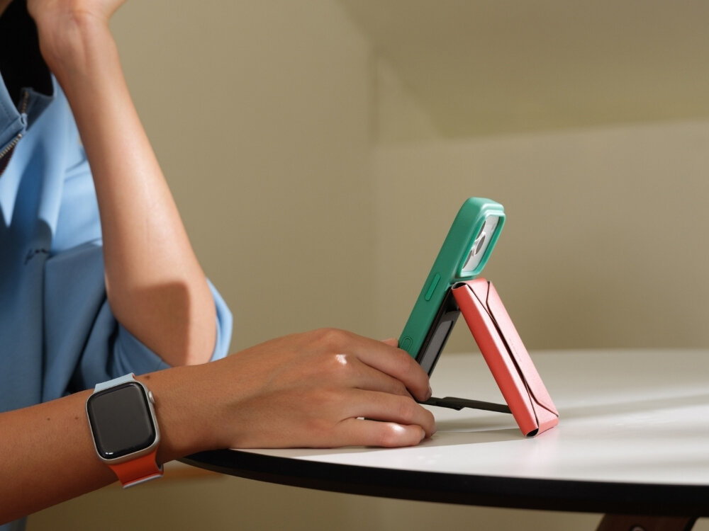 Osoba trzyma telefon w zielonym etui, który jest podparty bordowym stojakiem na białym stole. Na nadgarstku widoczny jest smartwatch z pomarańczowym paskiem.