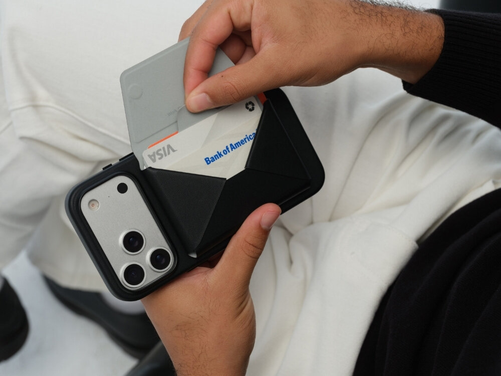Osoba trzyma czarny portfel MagSafe, do którego wkłada kartę z napisem \'Bank of America\'. Obok portfela znajduje się telefon z trzema obiektywami aparatu.
