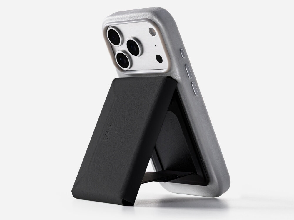 Czarny portfel MOFT Agile Travel MagSafe przymocowany do tylnej części telefonu w szarej obudowie. Portfel jest ustawiony pod kątem, pełniąc funkcję stojaka.