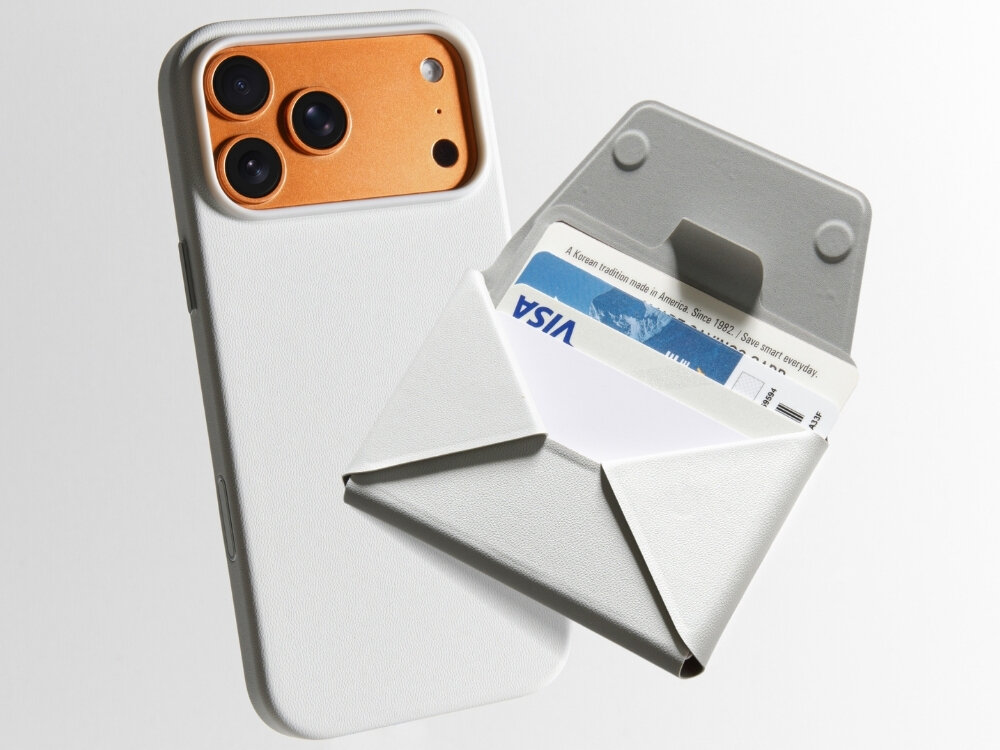 Na obrazie widoczny jest telefon z trzema aparatami w pomarańczowej obudowie oraz szary portfel MagSafe z kartą płatniczą z logo VISA. Portfel jest otwarty, ukazując miejsce na karty.